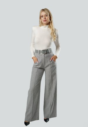 Blondynka stoi z rękami w kieszeniach, ubrana w biały przylegający golf, szare, szerokie spodnie z wysokim stanem, pasek oraz czarne, spiczaste buty.