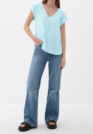 Woman wearing light blue V-neck short-sleeve top, ripped wide-leg jeans, and black platform sandals, standing with one hand in pocket.
