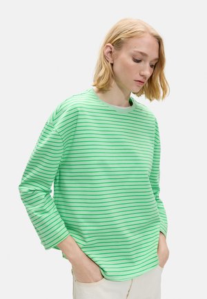 Młoda osoba o blond włosach do ramion, ubrana w zielony i jasnozielony paskowany top z długim rękawem, ręce w kieszeniach, patrzy w dół.