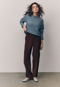 Maglione azzurro chiaro con scollo rotondo, abbinato a pantaloni marroni a gamba larga e scarpe beige. L'outfit presenta tessuti morbidi e una vestibilità rilassata.