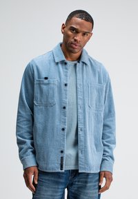 Man wearing light blue corduroy button-up shirt over blue t-shirt and jeans, standing against a plain light gray background.