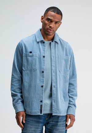 Man wearing light blue corduroy button-up shirt over blue t-shirt and jeans, standing against a plain light gray background.