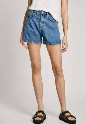 Kvinde iført blå denimshorts, hvid t-shirt delvist stukket ind, og sorte nittede sandaler, der står mod en ensfarvet baggrund.