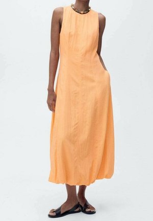 Długa, pomarańczowa sukienka bez rękawów o luźnym kroju, z lekkiego materiału i zaokrąglonym dekoltem. Modelka nosi czarne sandały i koralikowy naszyjnik.