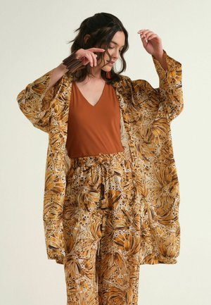 Woman wearing loose floral patterned pants and a matching long open jacket over a rust-colored V-neck top, adjusting her hair with raised arms.
