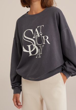 Femme portant un sweat-shirt gris foncé avec un grand texte blanc stylisé indiquant « SATURDAY » et un pantalon beige clair, une main dans la poche.