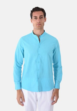 Uomo con camicia azzurro chiaro con bottoni e colletto alla coreana e pantaloni bianchi, in piedi davanti a uno sfondo bianco uniforme.