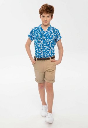 Blauw kortemouwen shirt met wit loofpatroon, beige opgerolde shorts, bruine gevlochten riem en witte sneakers. Eenvoudige, casual zomeroutfit.