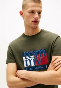Camiseta verde oliva de algodón, con un gran logotipo gráfico en azul, rojo y blanco. Mangas cortas y cuello redondo.