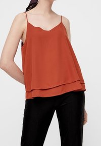 Rdzawoczerwony bezrękawnik z falbaniastym dekoltem, warstwowym fasonem, lekkim materiałem i cienkimi regulowanymi ramiączkami. W połączeniu z czarnymi spodniami.