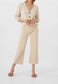 Kobieta w kremowym kardiganie z grubym splotem i dużymi guzikami, beżowych szerokich, skróconych spodniach oraz brązowych, plecionych sandałach na obcasie z odkrytymi palcami.