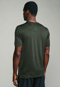 Dark green, short-sleeve athletic shirt with a textured surface and side slits; features a subtle reflective detail on the back neckline.