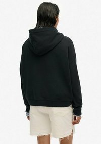 Felpa nera con cappuccio e vestibilità rilassata. Tessuto in misto cotone, con polsini e orlo a costine. Indossata sopra pantaloni corti bianchi con bordi sfrangiati.