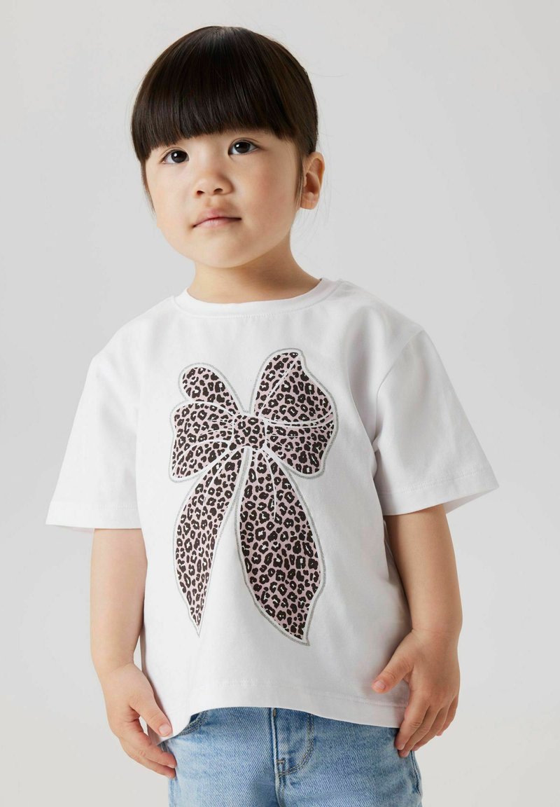 Camiseta de algodón blanca con un gran diseño de lazo de estampado de leopardo con acentos rosas. Mangas cortas y un corte relajado, combinada con jeans azul.