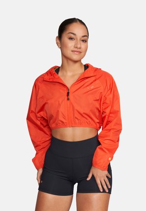 Młoda kobieta z włosami związanymi do tyłu, ubrana w krótki, jaskrawo pomarańczowy, zapinany na zamek krótki płaszcz oraz wysokie, czarne, sportowe szorty, stojąca na tle jednolitego tła.