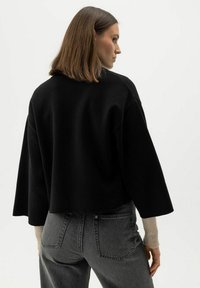 Kvinna med axellångt brunt hår bär en lös svart topp över beige långärmade tröja och högmidjade mörkgrå jeans, vänd åt sidan.