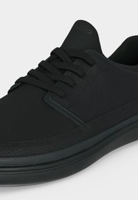 Gros plan d'une basket noire avec des panneaux latéraux texturés, des lacets noirs et une semelle noire lisse.