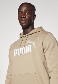 Beige Hoodie aus weichem Material mit großem weißen "PUMA"-Logo und einem springenden Katzen-Graphic auf der linken Brustseite. Mit einer Fronttasche.