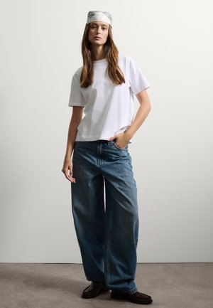 T-shirt bianco a maniche corte abbinato a jeans blu a gamba larga, con un leggero foulard a fantasia e mocassini scuri su uno sfondo neutro.