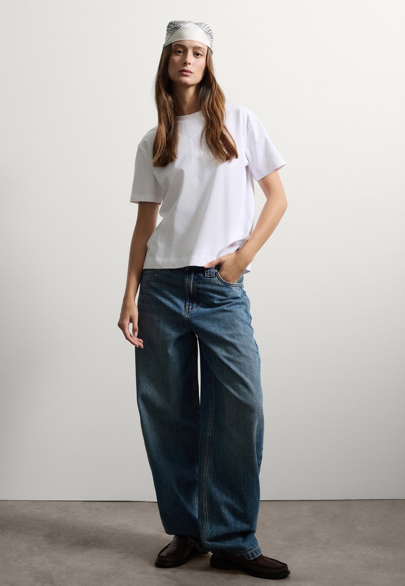 Camiseta blanca de manga corta combinada con vaqueros de pierna ancha azules, con un pañuelo ligero de estampado y mocasines oscuros sobre un fondo neutro.