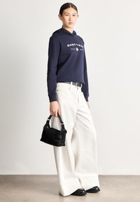 Námořnická mikina s kapucí s nápisem "GANT 1949", volné bílé široké kalhoty, černá kožená taška a robustní černé boty. Hladké textury a moderní design.