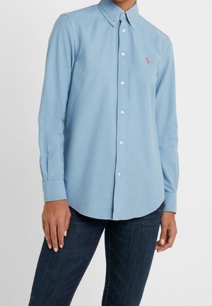 Person trägt ein hellblaues Hemd mit Knöpfen und Kragen, kombiniert mit dunkelblauer Jeans, vor einem schlichten weißen Hintergrund.