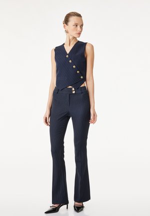 Femme portant un gilet sans manches bleu marine à fines rayures avec des boutons dorés et un pantalon évasé assorti, associé à des escarpins noirs pointus.