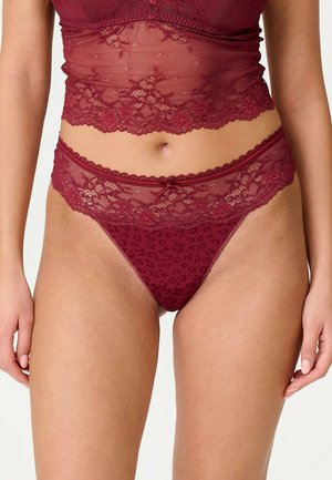 Femme portant un ensemble de lingerie en dentelle bordeaux avec des motifs floraux, montrant le ventre et le haut des cuisses sur un fond neutre.