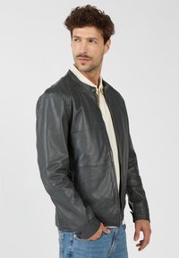 Donkergrijze leren jas met een staande kraag, rits aan de voorkant en zakken aan de zijkanten. Gedragen over een lichtgekleurde shirt. Gladde textuur.