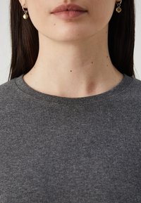 Maglione grigio a coste con scollo rotondo, caratterizzato da una texture morbida. Orecchini piccoli e rotondi in argento con un accento beige.