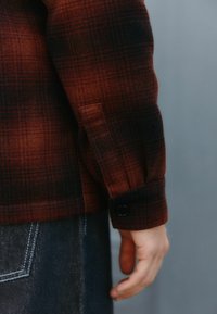 Ärmel einer rot-schwarzen karrierten Jacke mit Knopfdetails, kombiniert mit einem Jeansstoff mit kontrastierenden Nähten. Texturiertes Wollmischmaterial.