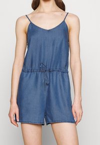 Jeansowy kombinezon z dekoltem w kształcie litery V, cienkimi regulowanymi ramiączkami, elastycznym pasem z wiązaniem oraz luźnym krojem z szortami.