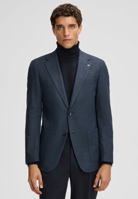 Man in een donkerblauwe geblokte blazer over een zwarte coltrui, staande tegen een effen lichtgrijze achtergrond.