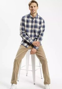 Koszula w kratkę z guzikami w niebiesko-białe paski, z długimi rękawami i kołnierzykiem. W połączeniu z beżowymi spodniami i białymi trampkami. Siedzi na stołku.