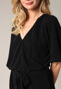 Robe noire style portefeuille avec des manches courtes, fabriquée en tissu texturé avec des fibres métalliques. Comprend un lien à la taille pour un détail supplémentaire.