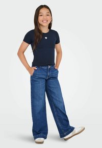 Fille souriante avec de longs cheveux noirs portant un haut noir à manches courtes avec un détail en forme de cœur, un jean large bleu, des baskets blanches, les mains dans les poches.