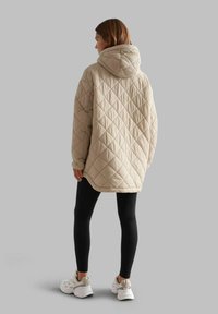 Beige quilted jacket with a hood, featuring a diamond pattern. Model wears black leggings and white sneakers, standing against a gray background.