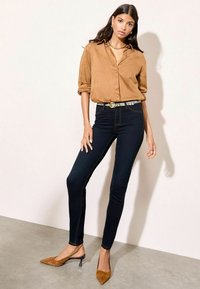 Camisa de ante marrón con botones, jeans oscuros ajustados con costuras en contraste y tacones claros de punta marrón. Cinturón de patrón sutil.