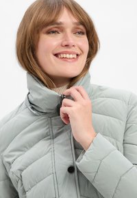 Hellgraue gepolsterte Jacke mit hohem Kragen. Mit Reißverschluss und schwarzen Knopfdetails ausgestattet, gefertigt aus einem glatten, gesteppten Material.