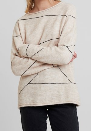 Persona con un suéter beige con finas líneas geométricas negras y pantalones negros, con los brazos cruzados, sobre un fondo gris claro liso.