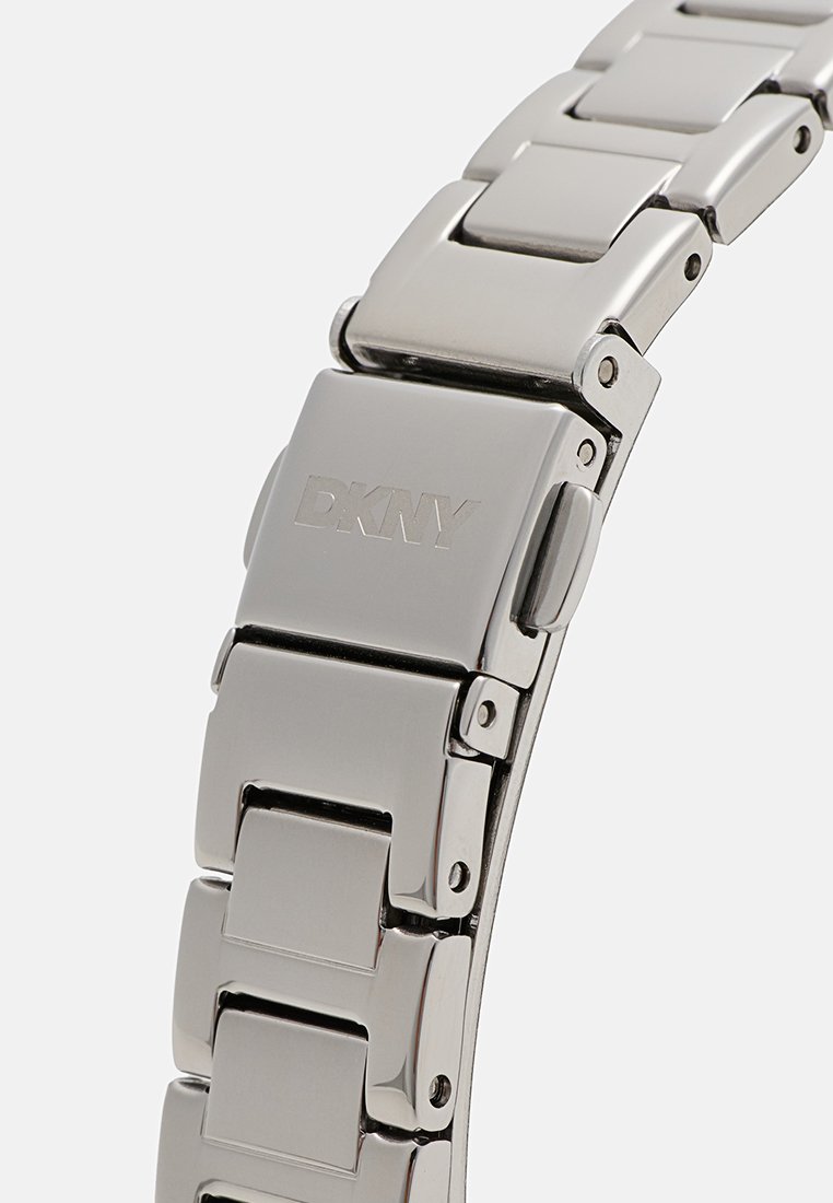 Pulsera de reloj de acero inoxidable plateado con eslabones rectangulares, superficie pulida y logo de DKNY grabado en el cierre. Diseño duradero y elegante.