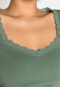 Top verde con scollatura in pizzo a onde, caratterizzato da dettagli delicati. Il tessuto è morbido, liscio e aderente nel design.