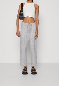 Kvinna i vit croppad tanktopp, ljusgrå drängsbyxor, svarta sandaler och bär en svart axelväska mot en enkel bakgrund.