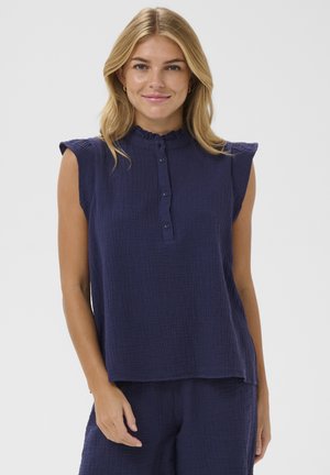 Žena s blond vlasmi sa usmieva, má na sebe námornícky modrý textúrovaný top bez rukávov s volánovým golierom a zapínaním na gombíky, v kombinácii s rovnakými námornícky modrými nohavicami.