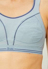 Tuva persona, kas valkā gaiši zilu sporta krūšturi ar tumši zilu apdari un "Champion Shock Absorber" uzlīmi joslā.