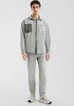 Lichtgrijze denim shirt met een donkergrijze patch, gecombineerd met lichtgrijze jeans en grijze sneakers. Beschikt over een knoopsluiting en een borstzak.