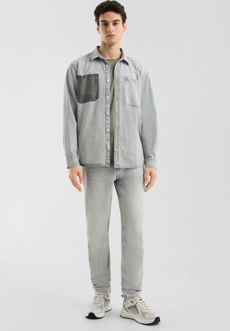 Helles graues Denimhemd mit einem dunkelgrauen Patch, kombiniert mit hellgrauen Jeans und grauen Sneakers. Verfügt über ein Button-Up-Design und eine Brusttasche.