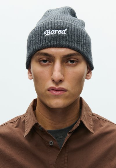 Giovane uomo con espressione neutra, che indossa un berretto di maglia grigio con l'etichetta "Bored" e una camicia marrone abbottonata sopra una maglietta scura.
