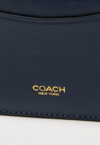 Πορτοφόλι από δέρμα σε ναυτικό μπλε χρώμα με απαλή υφή και ανάγλυφο λογότυπο "COACH NEW YORK" σε χρυσό. Τακτικές ραφές κατά μήκος των άκρων.