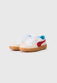 Białe i beżowe sneakersy z czerwonymi akcentami, z cholewką zamszową i tekstylną, niebieskim wnętrzem i gumową podeszwą. Klasyczny wzór sznurowania.